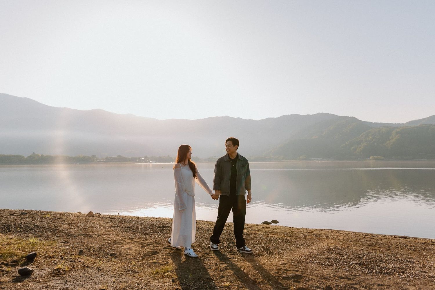 Couple holding hands by a serene lake with misty mountains and golden sunlight in the background.