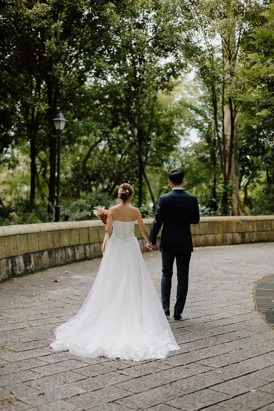 Bride in flowing white dress and groom walk along curved stone path surrounded by green trees.