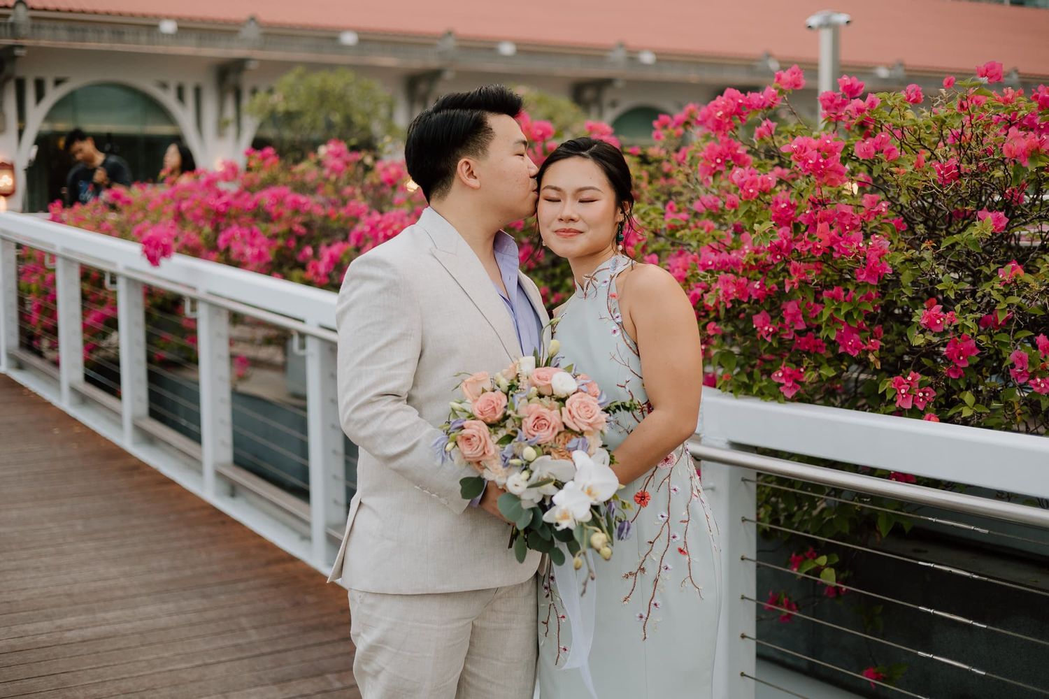 A couple poses with a pink bouquet against blooming bougainvillea on an outdoor deck.