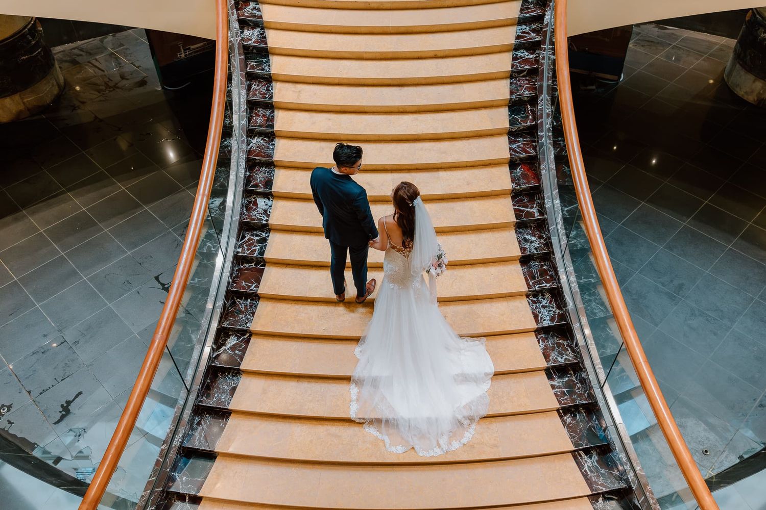 Wedding party ascends curved wooden staircase with ornate black metal railings.