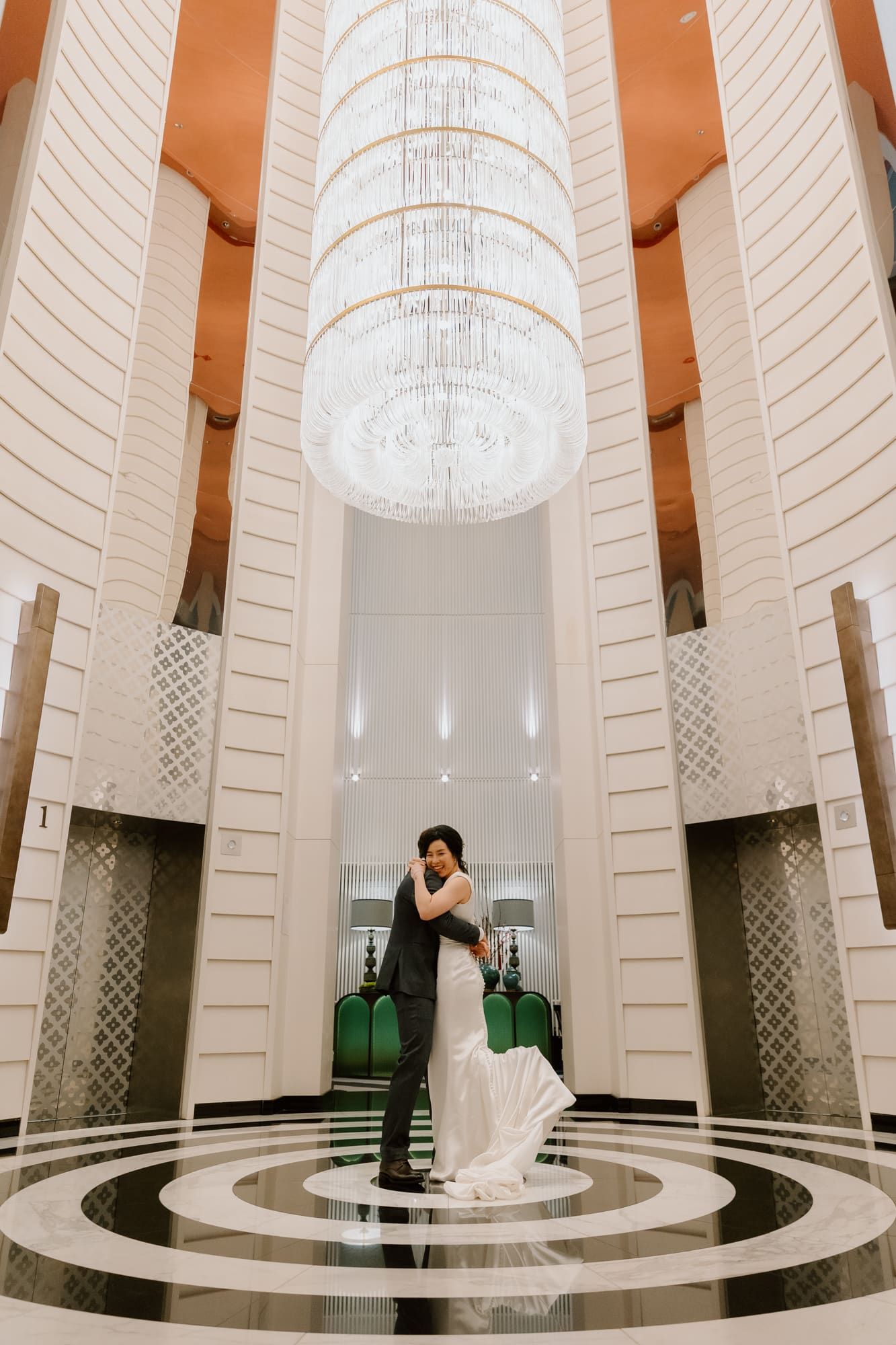 A bride poses in a stunning white gown inside an elegant modern white circular atrium with geometric patterns and chandeliers.