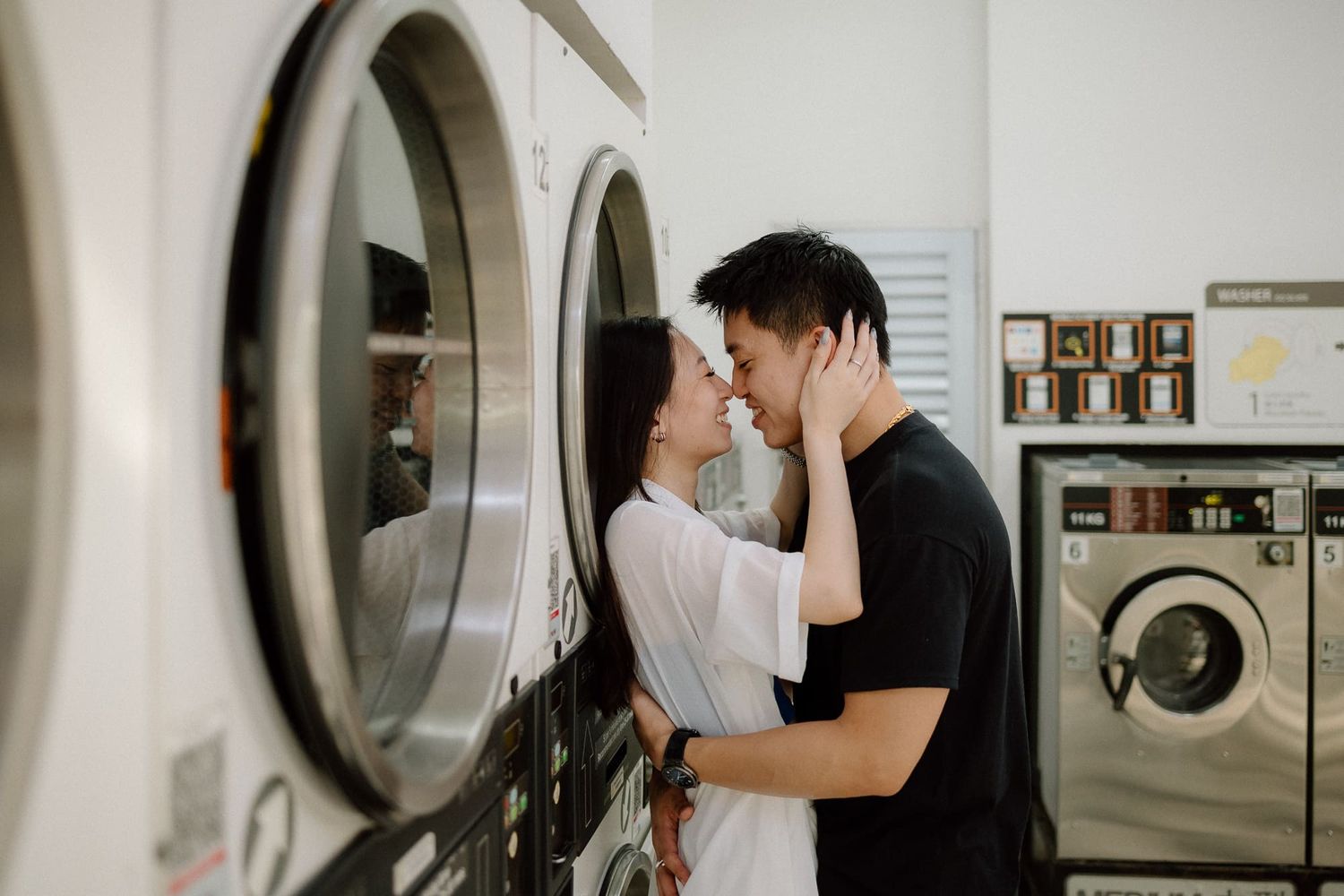 Romantic couple embraces near washing machines in a bright, minimalist laundromat setting.
