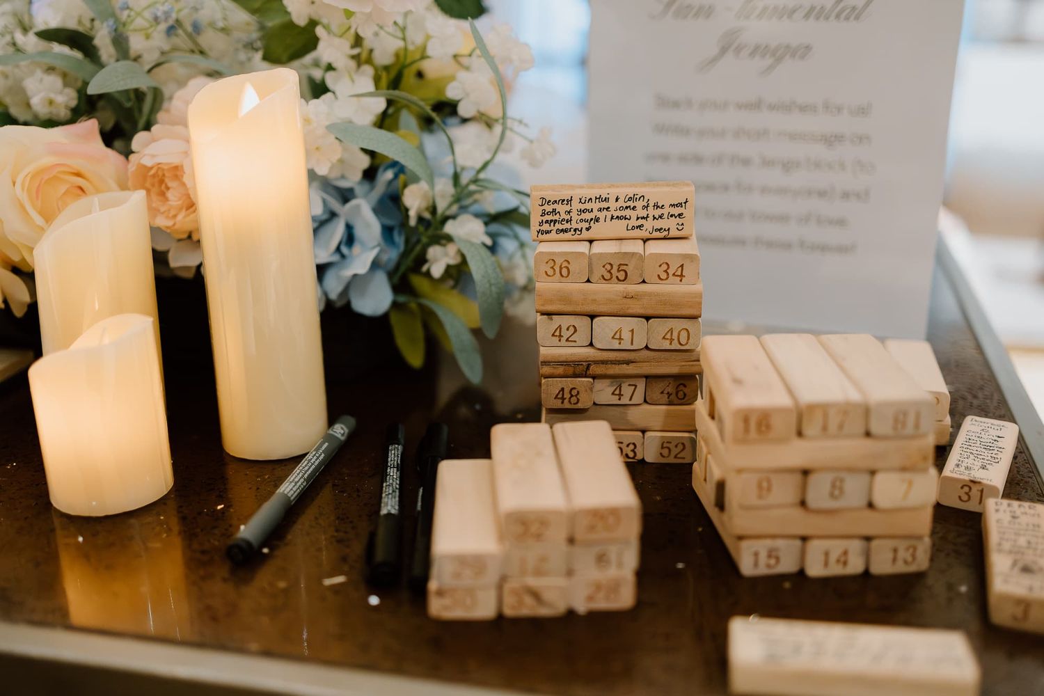 Wedding reception table with wooden Jenga blocks and glowing white candles beside floral arrangements.