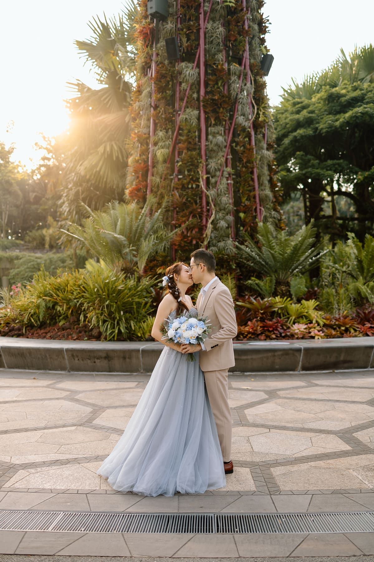 A romantic wedding portrait in a lush garden with palm trees and golden sunlight at sunset.