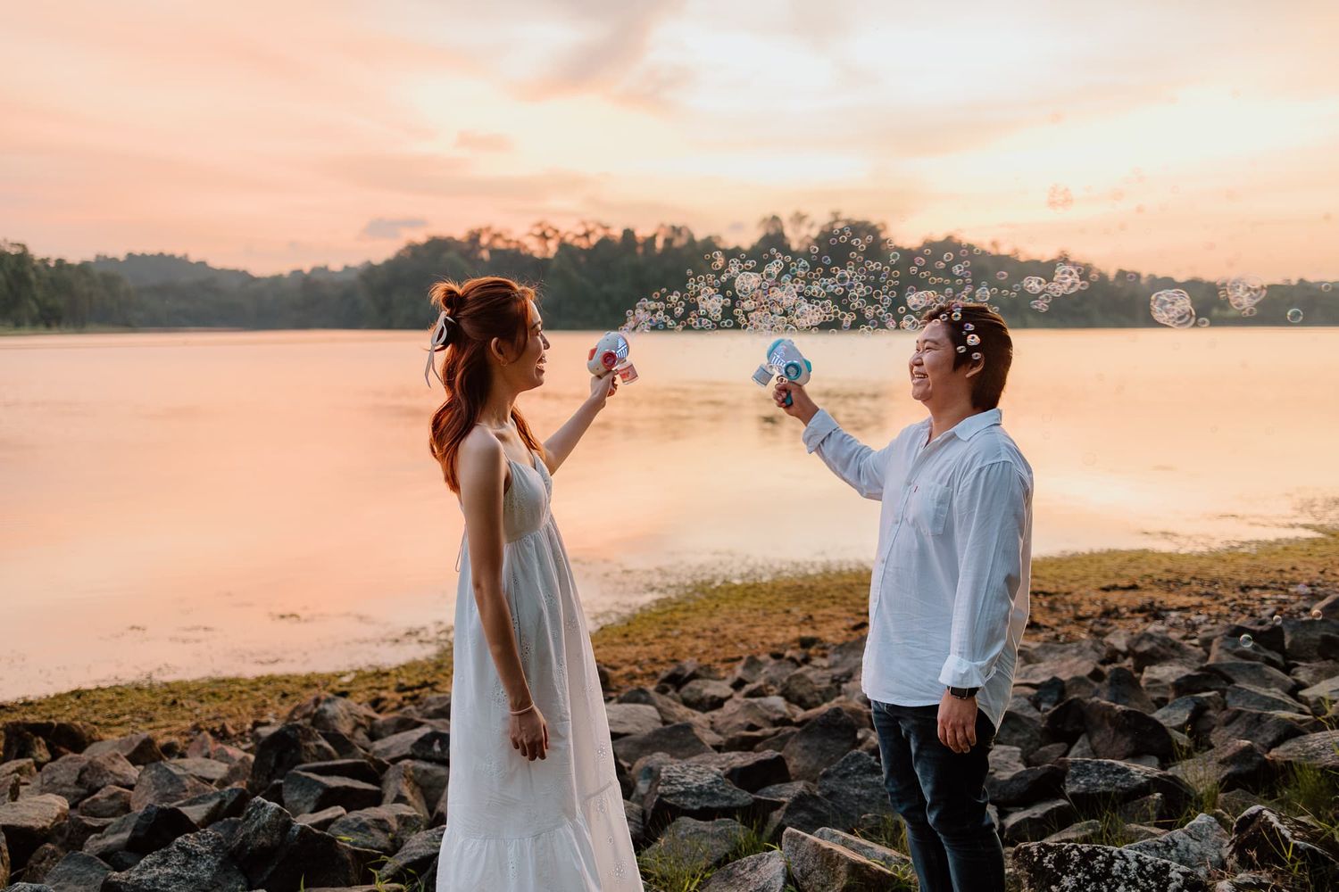 Couple in white outfits toasting by lakeside during golden sunset.
