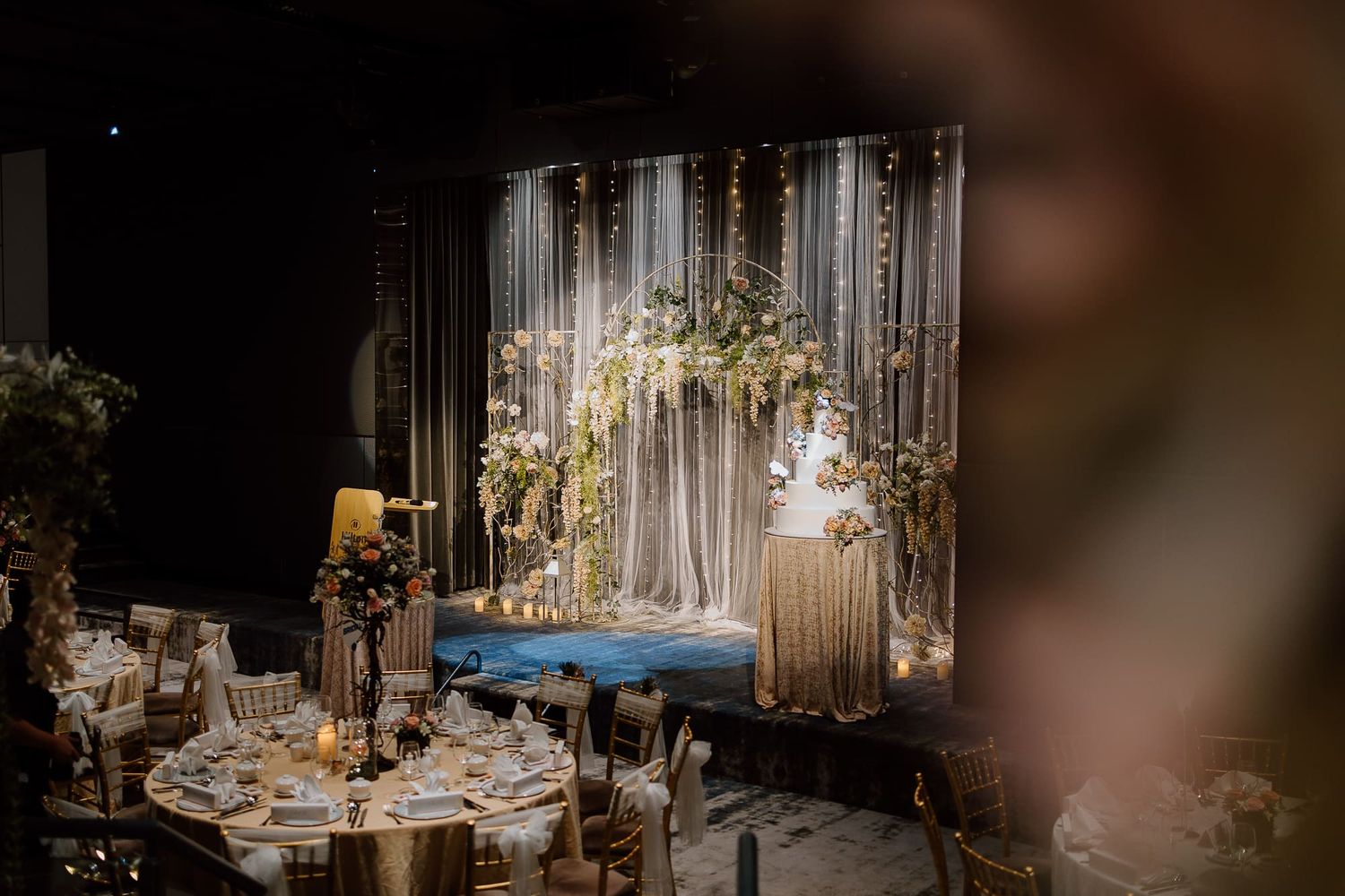Elegant wedding reception setup with white drapery backdrop, floral arrangements and round dining tables with glowing candles.