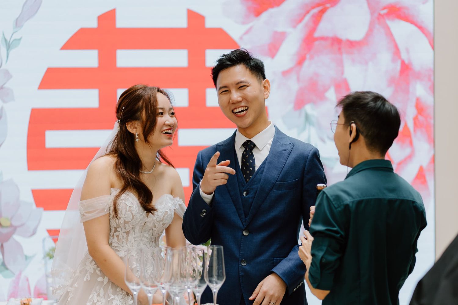 Wedding celebration scene with guests toasting in front of a decorative red double happiness symbol backdrop.