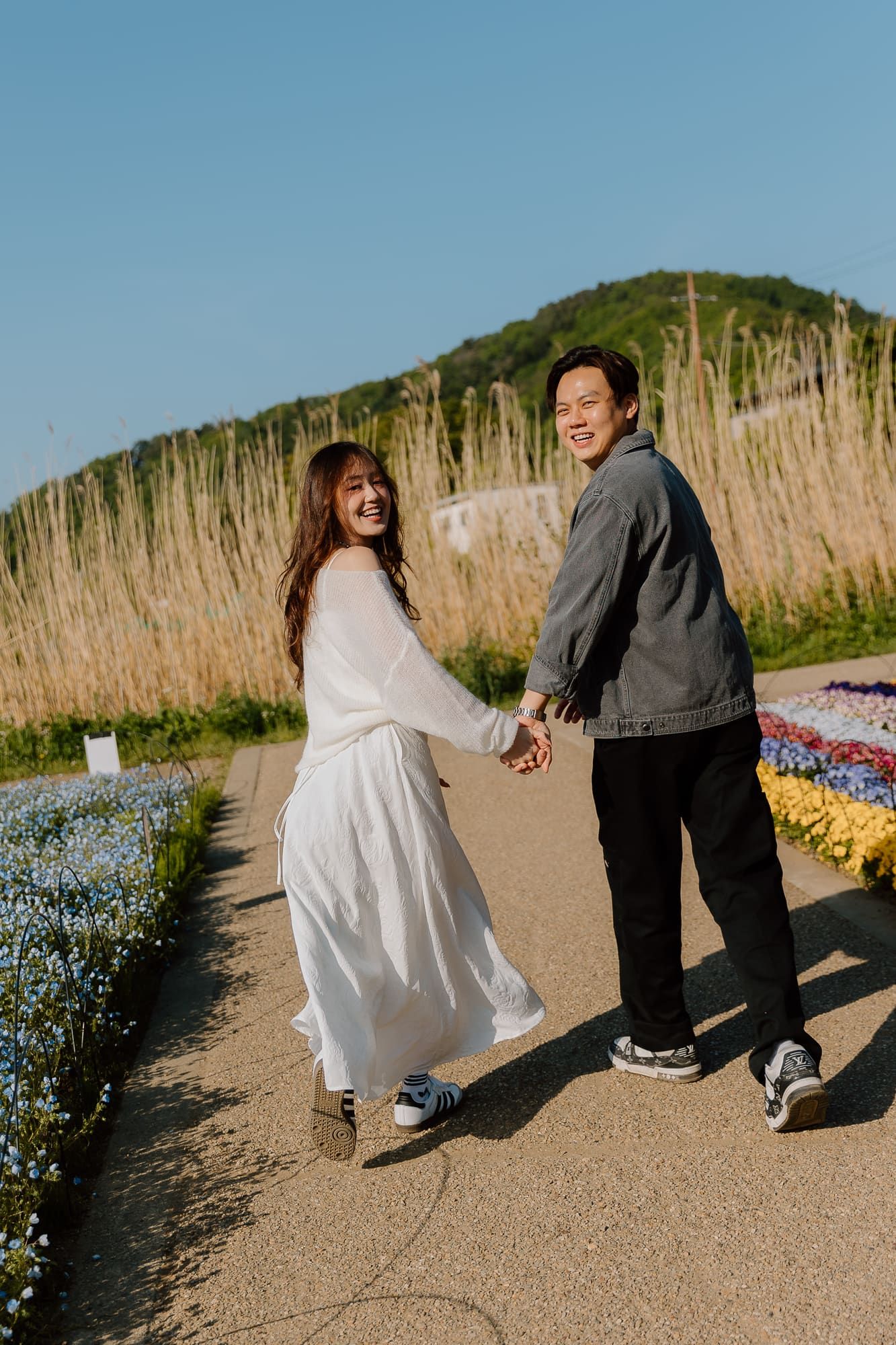 Happy couple holding hands on a flower-lined path with a green hill and blue sky backdrop.