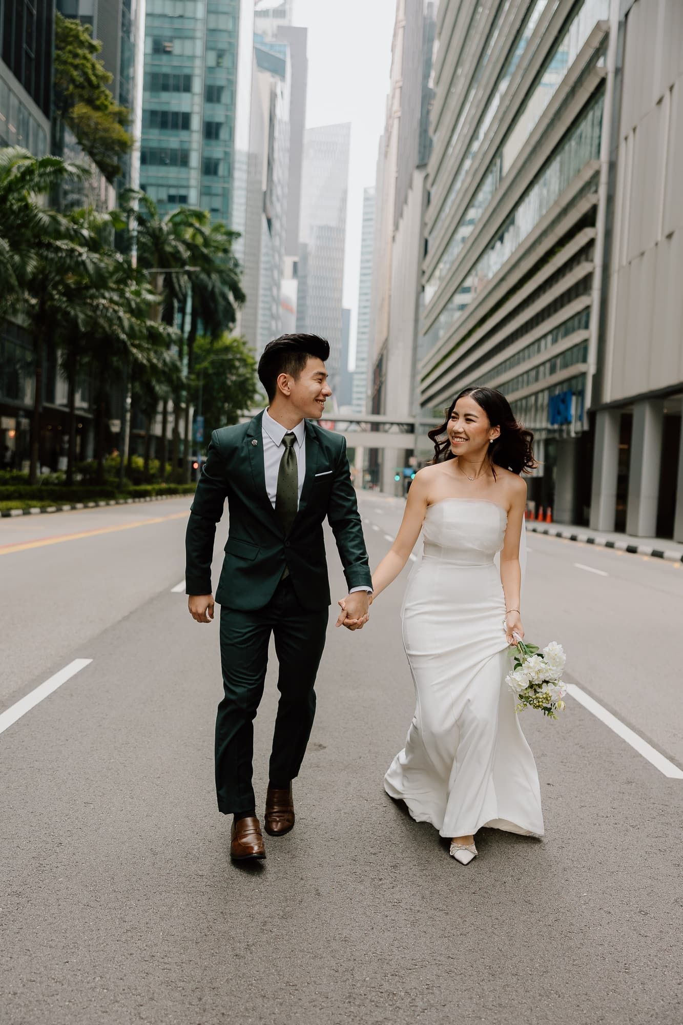 A bride and groom walk hand in hand down an empty city street surrounded by skyscrapers.