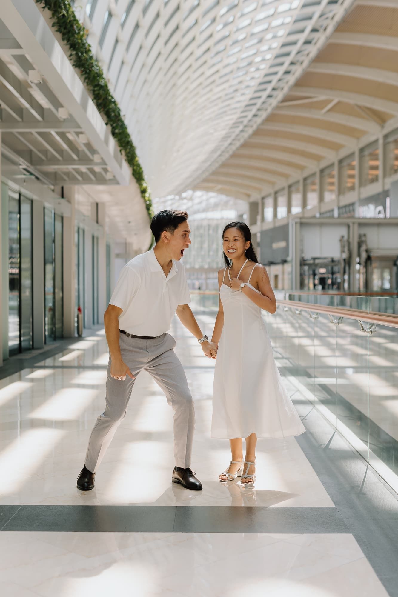 Couple in white and grey outfits walking through a modern bright hallway with architectural details.