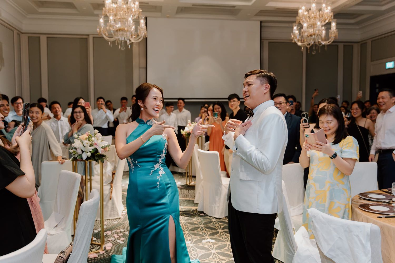 Guests in formal attire singing karaoke together at an elegant wedding reception with crystal chandeliers.