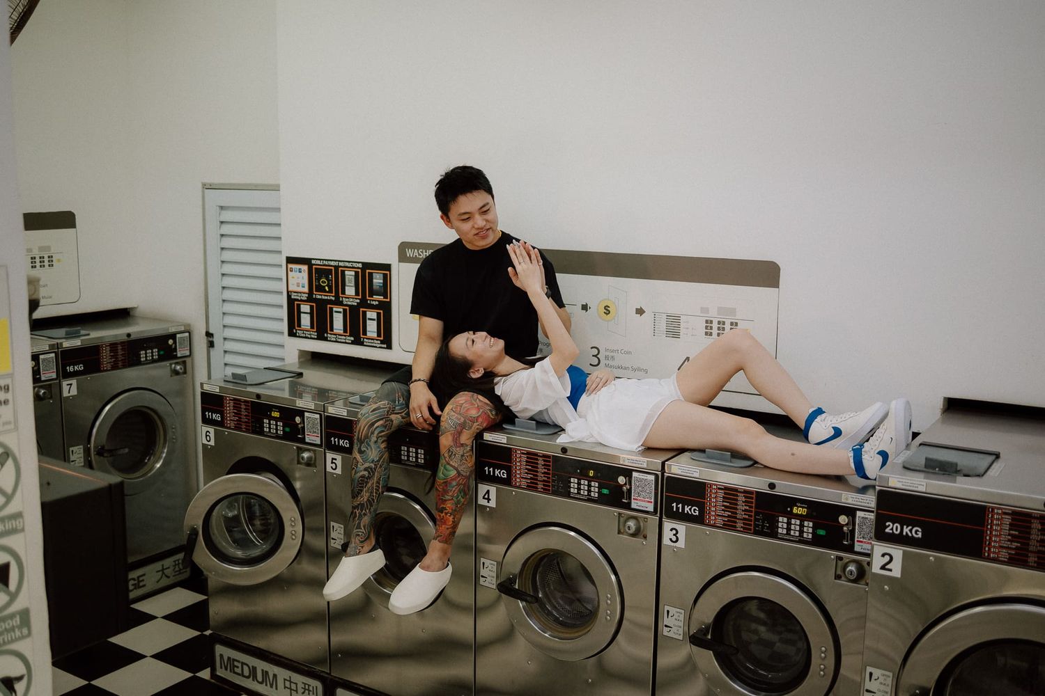 Two people share a playful moment while sitting on top of washing machines in a laundromat.