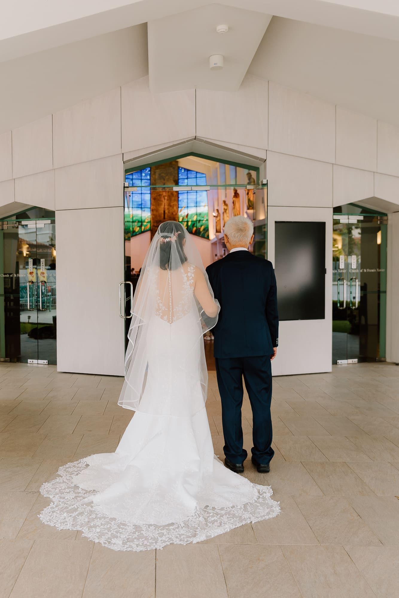 A bride and groom stand together in a church atrium with a flowing white lace wedding gown trailing behind them.