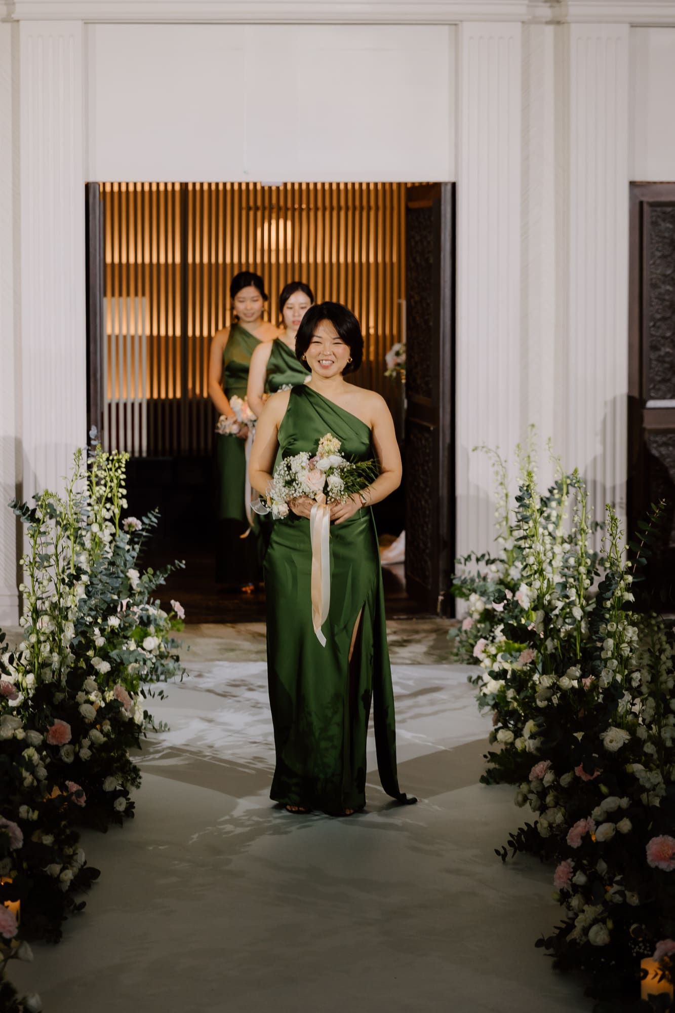 Bridesmaid in emerald green dress walks down floral-lined aisle at indoor wedding venue.
