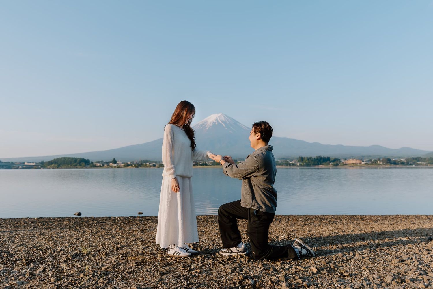 Man proposing to woman on rocky lakeside with Mount Fuji in background on clear day.