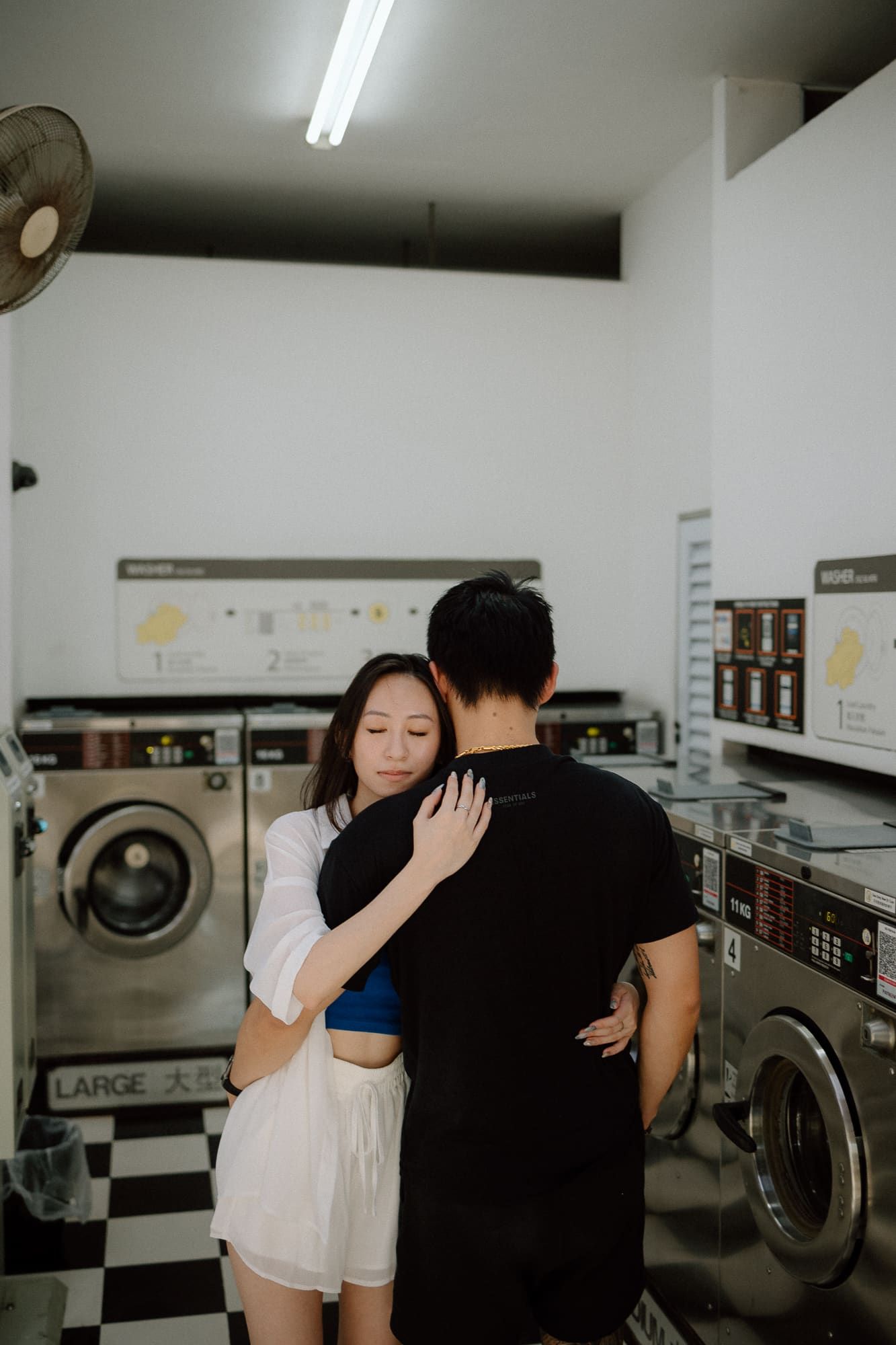 A couple embraces intimately in a laundromat with industrial washing machines in the background.