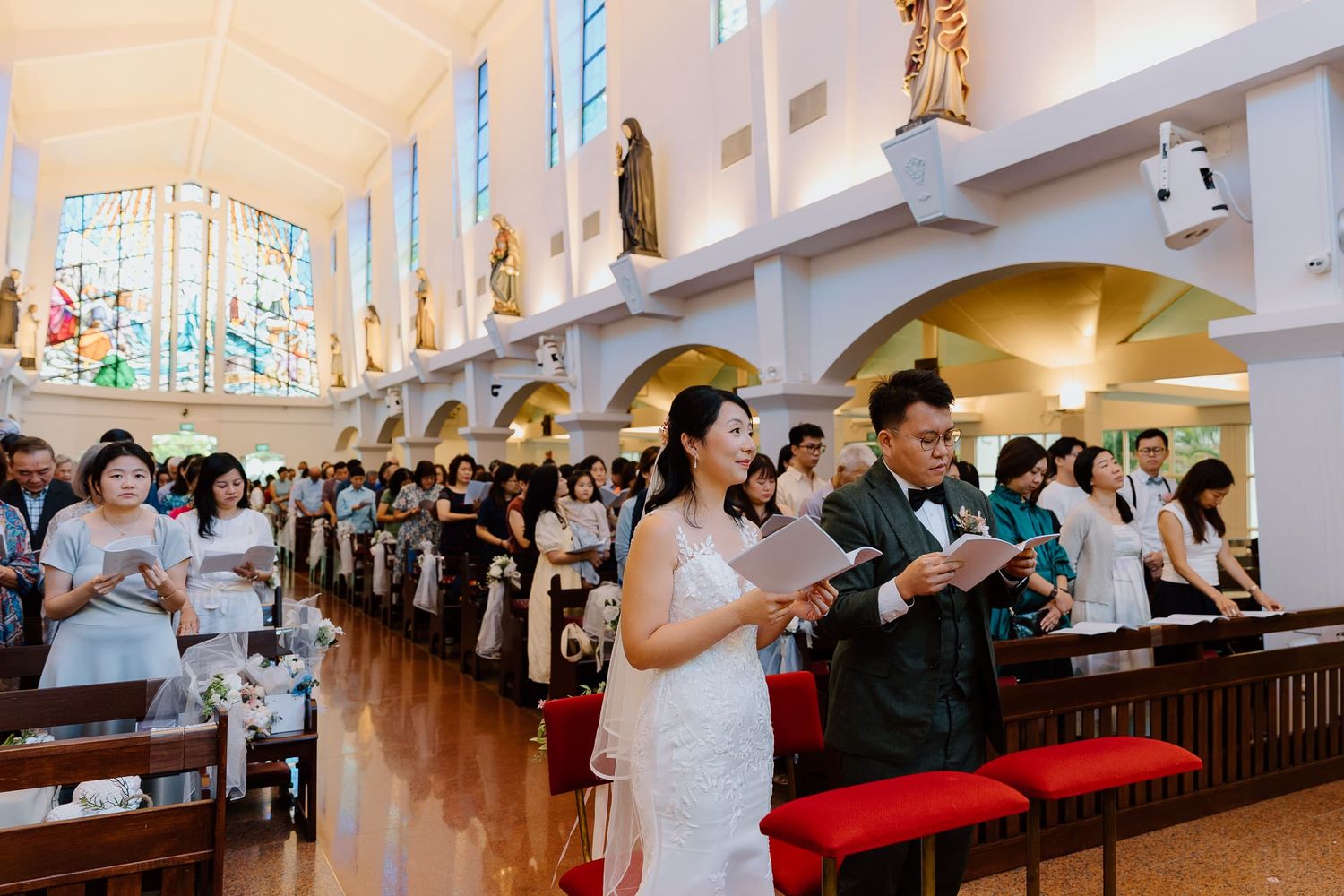 Catholic church congregation gathers for worship service in bright white chapel with stained glass windows and red kneelers.