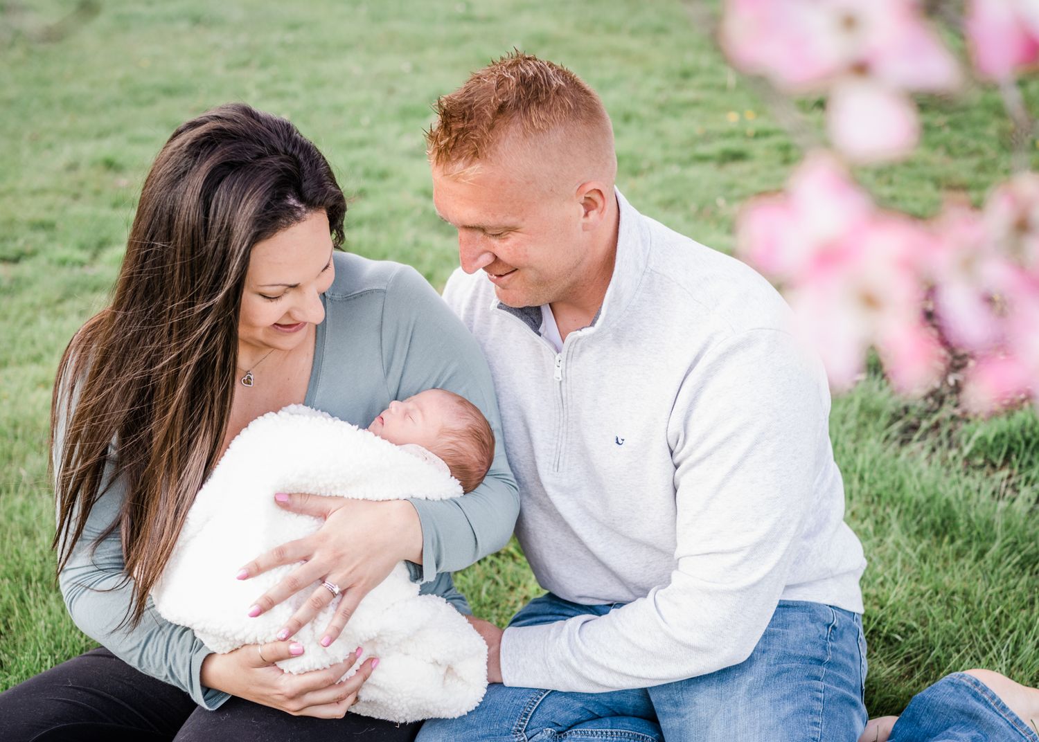 Family sits on grass with newborn baby during spring photo session with pink blossoms.
