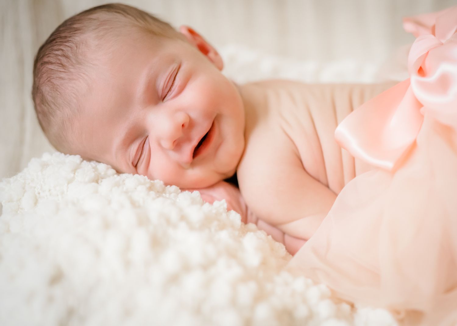 Peaceful newborn baby sleeping on fluffy white blanket with soft peach-colored lighting.