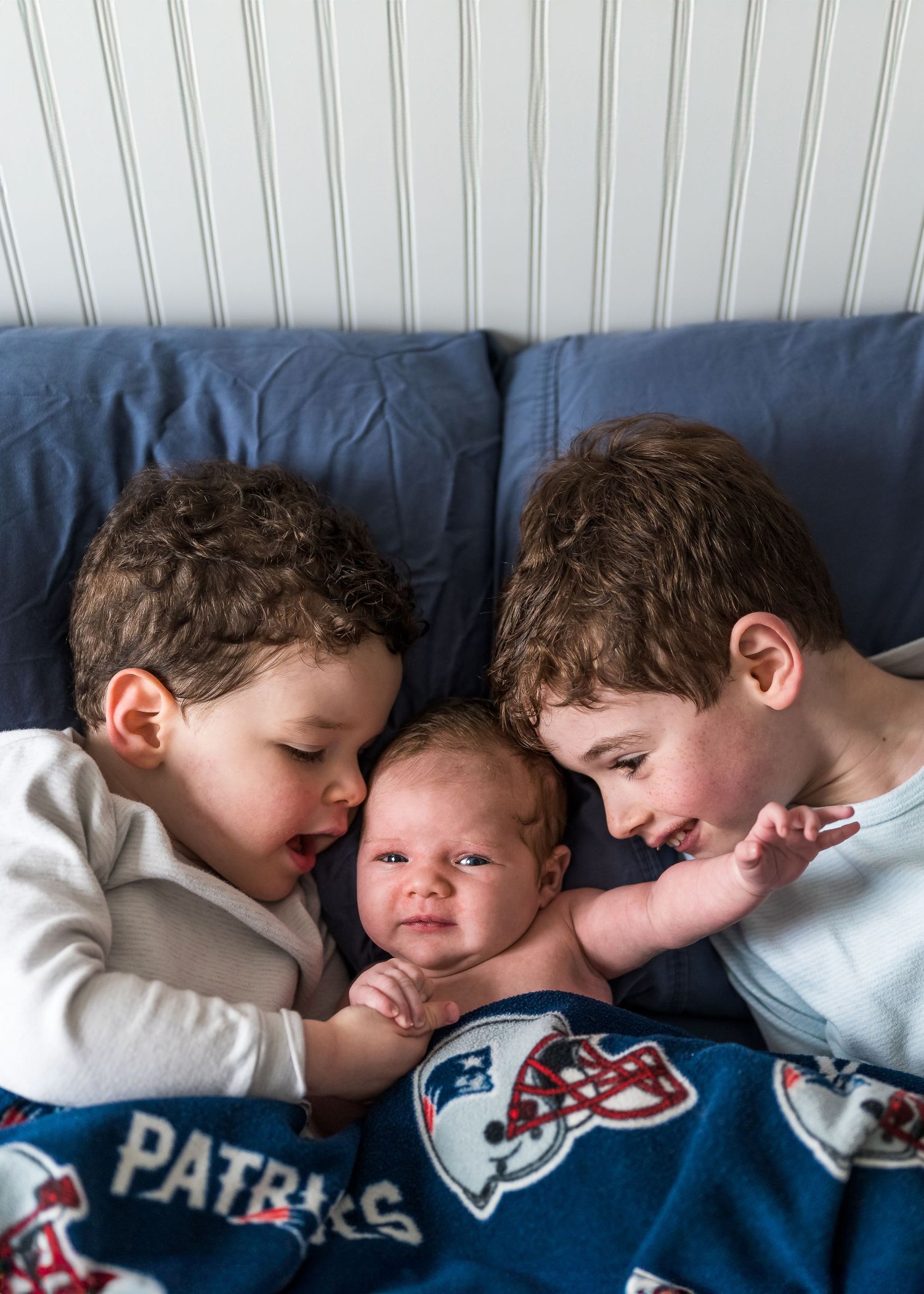 Three children cuddle together on a blue couch with a Patriots sports blanket.