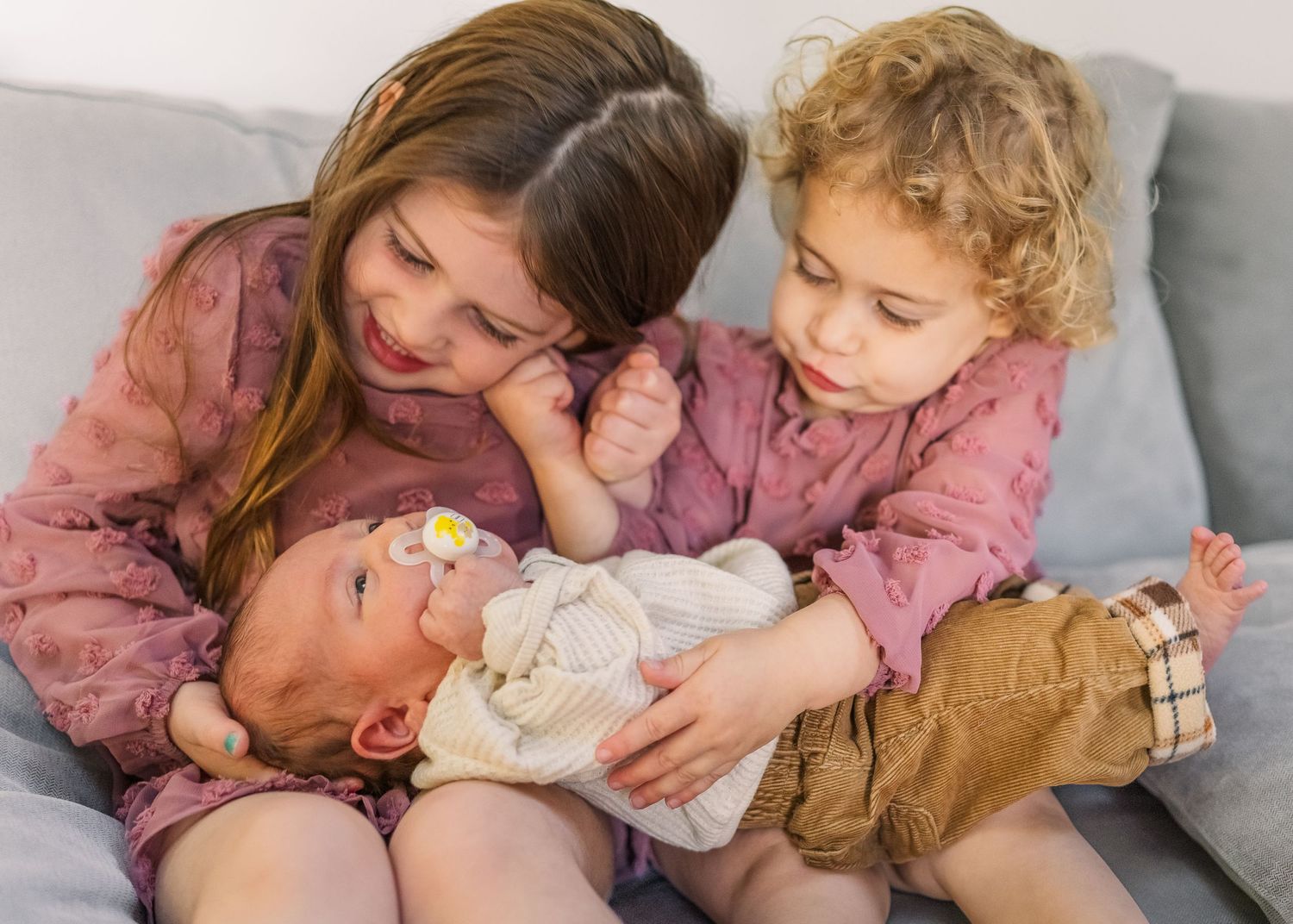 Two siblings in pink outfits bonding with their newborn baby sibling while sitting on a couch.