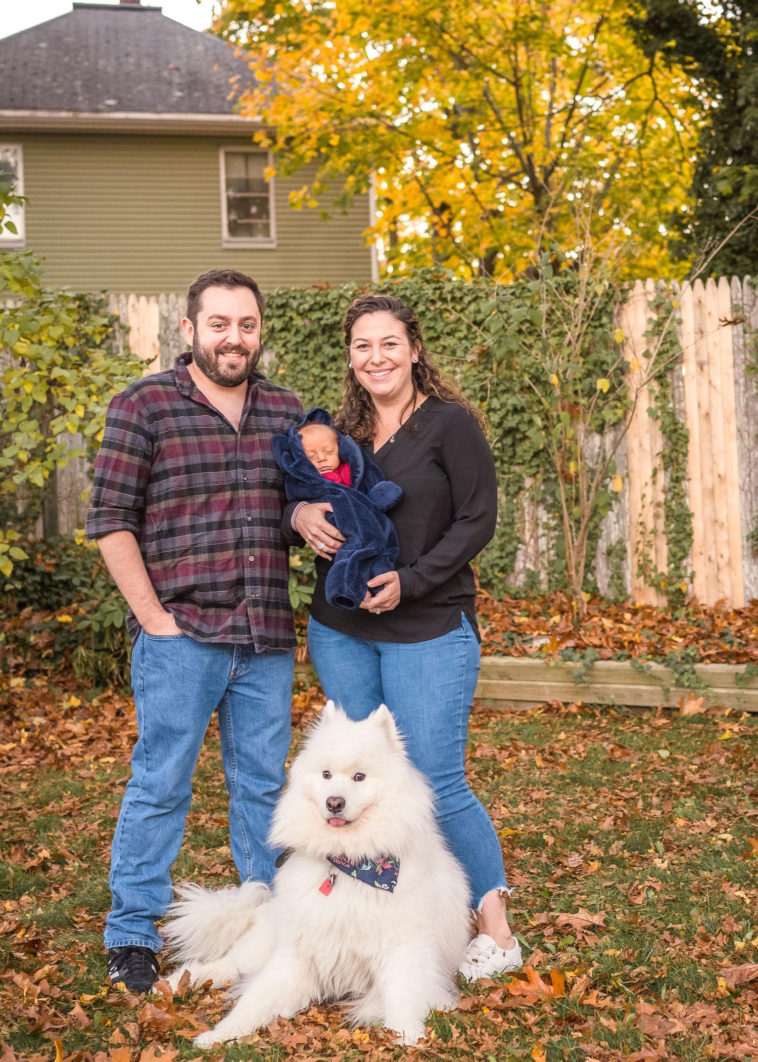 Family poses with their fluffy white dog in autumn backyard setting.