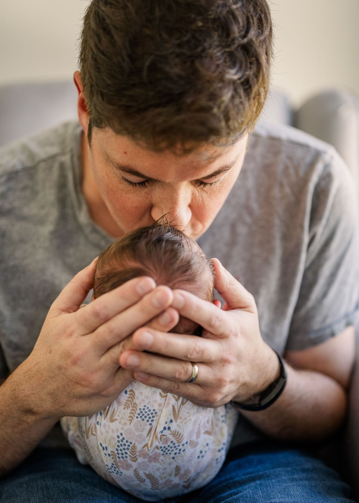A parent in a gray t-shirt tenderly kisses their infant child's head while cradling them in a floral pattern outfit.