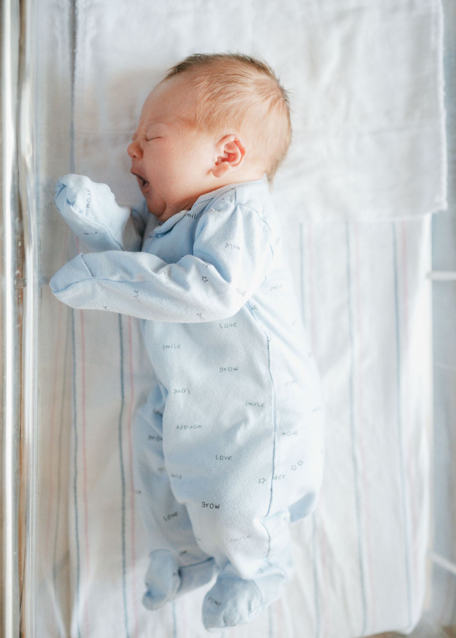 Newborn baby in a pale blue onesie sleeping peacefully on white bedding.