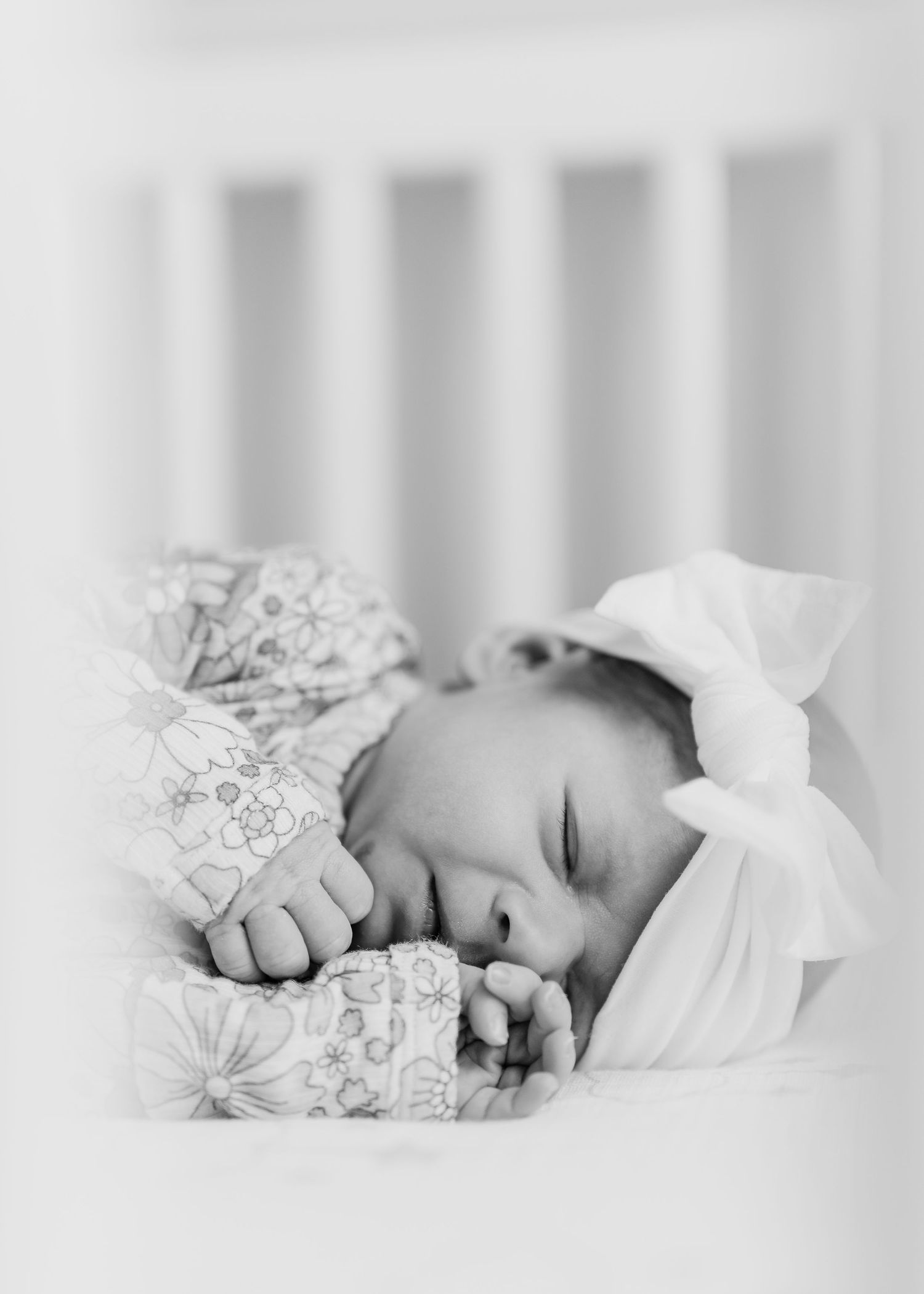 Sleeping baby in white bow headband viewed through crib slats in black and white.