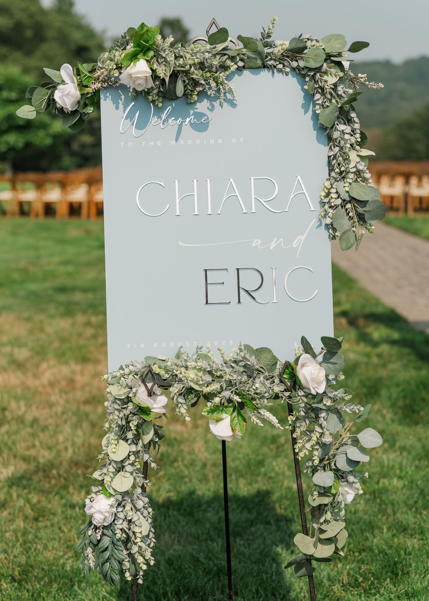 Elegant wedding welcome sign with floral eucalyptus garland decorations and names Chiara and Eric on light blue background.