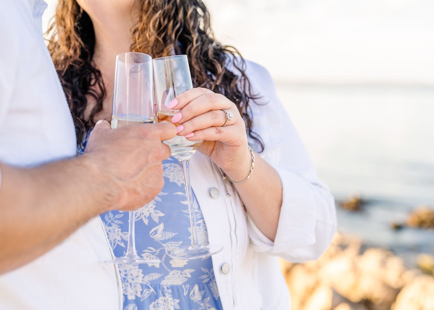 Two hands toasting with champagne glasses against a sunset beach background.