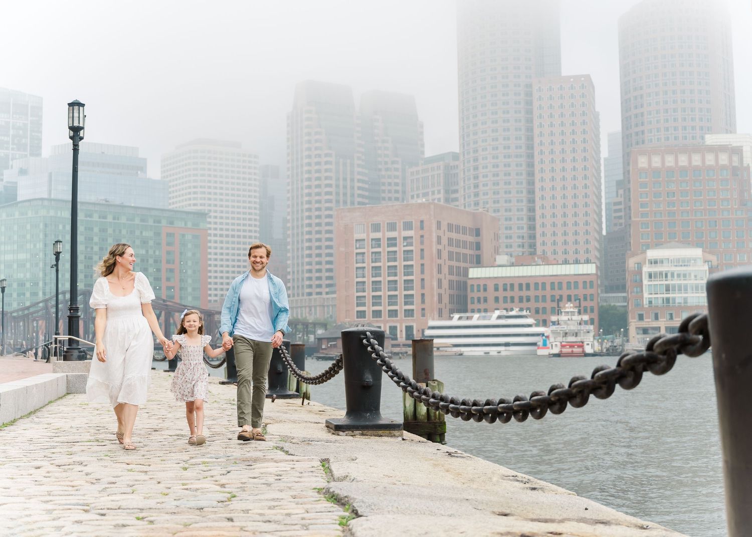 Family strolling along waterfront path with chain barrier and foggy cityscape backdrop.