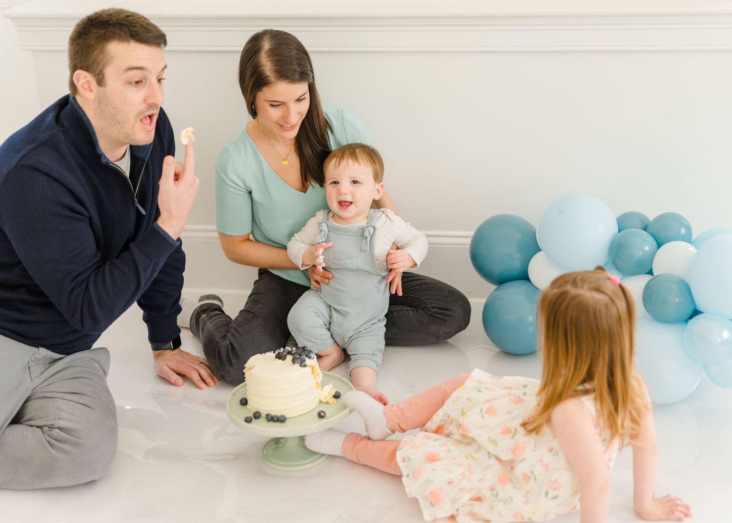 Family celebrating baby's first birthday with cake and blue balloons during an intimate party photoshoot.