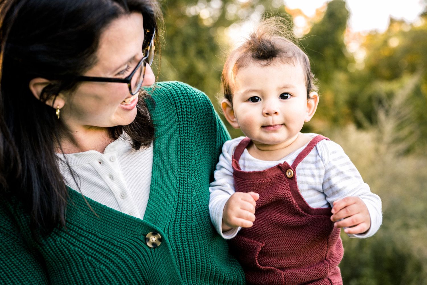 An intimate portrait of a parent in a green sweater holding a smiling baby in burgundy overalls.