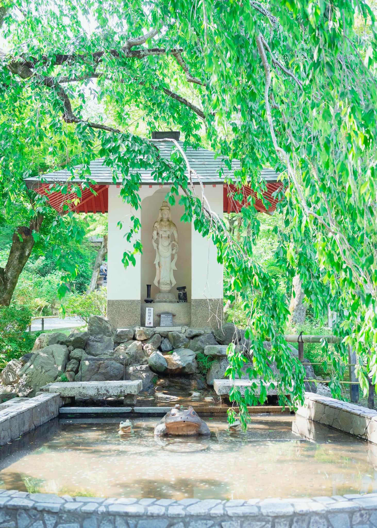 Small white shrine with statue surrounded by willow trees and stone landscaping.