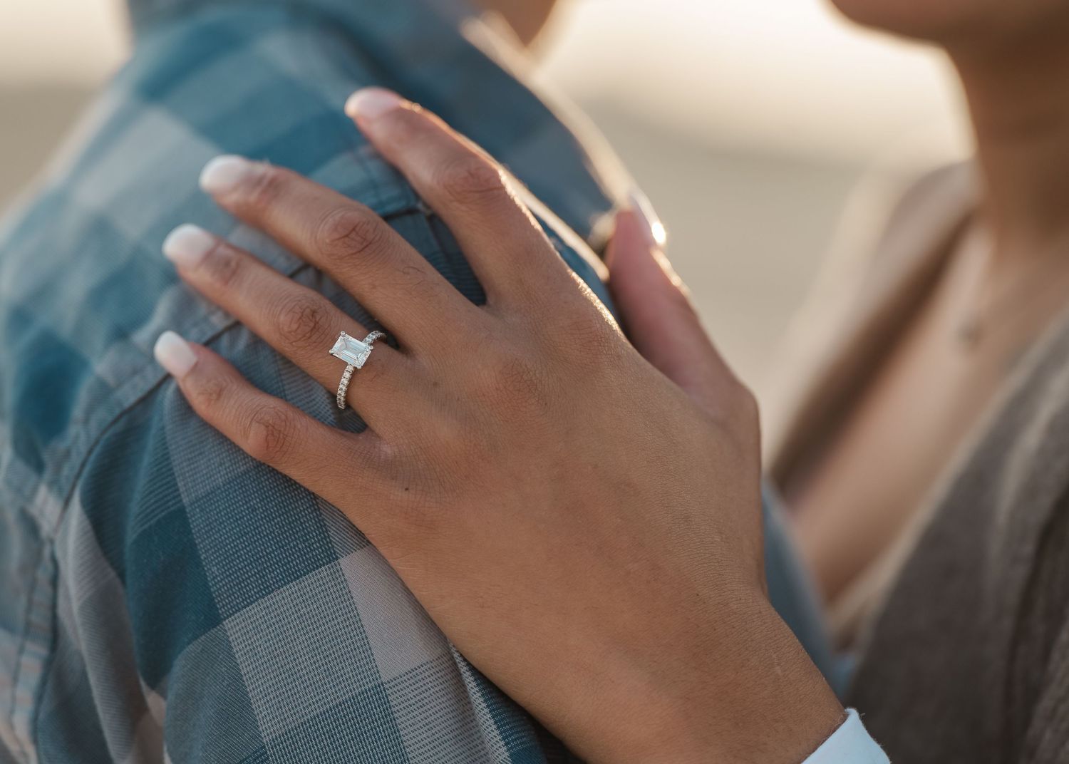 A hand wearing an engagement ring rests gently on turquoise plaid fabric in soft natural lighting.
