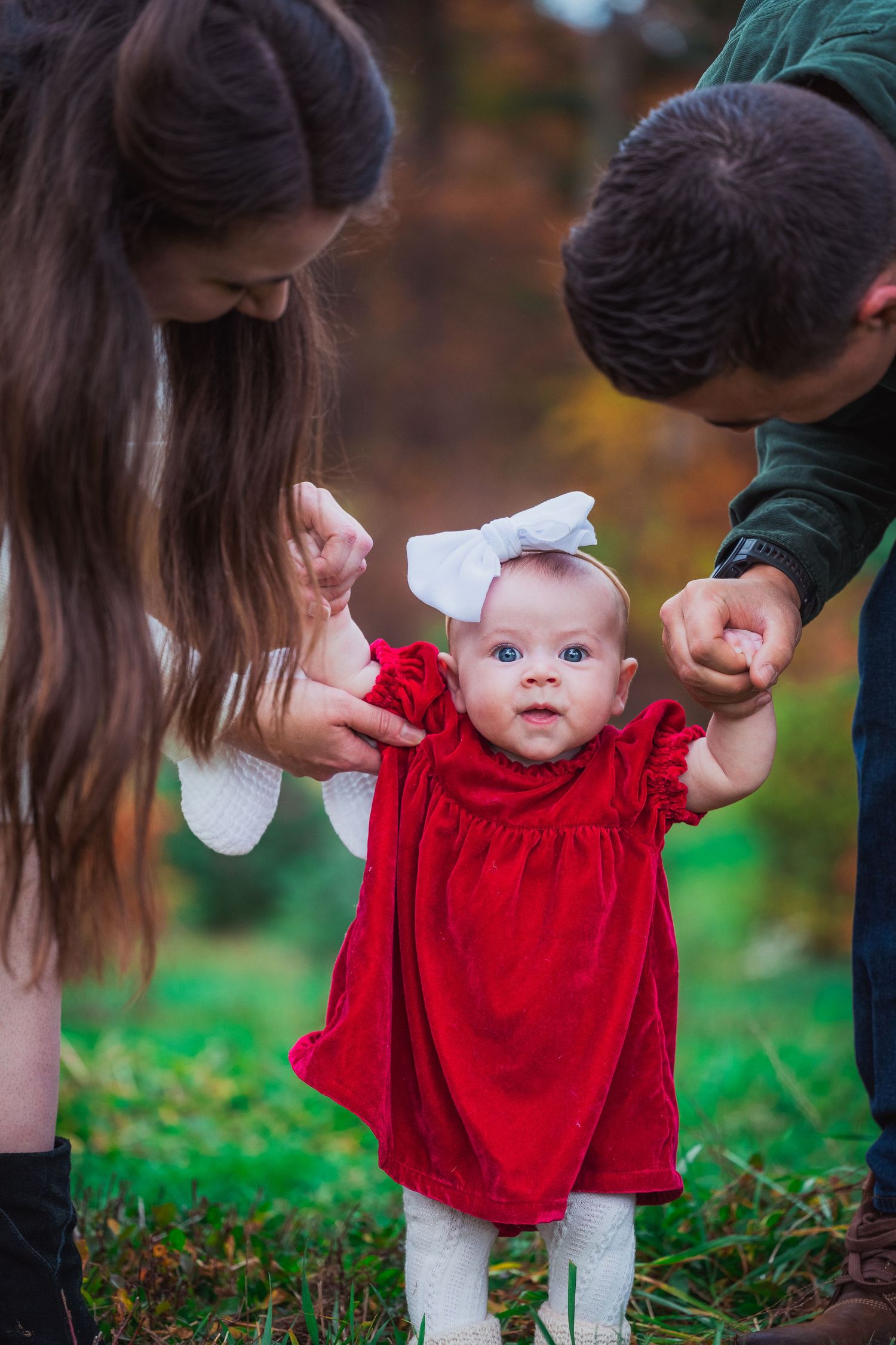 A baby in a red dress with white tights is supported while standing on grass during an autumn photo session.