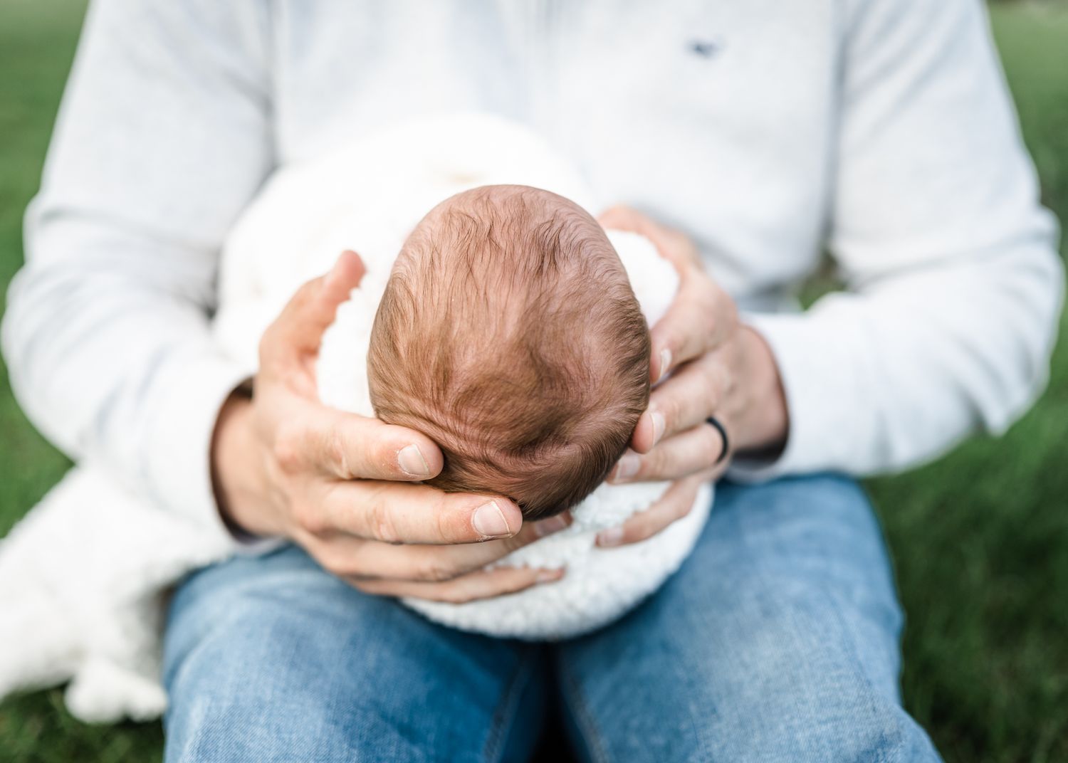 Close-up of hands gently cradling newborn wrapped in soft white blanket on grass.