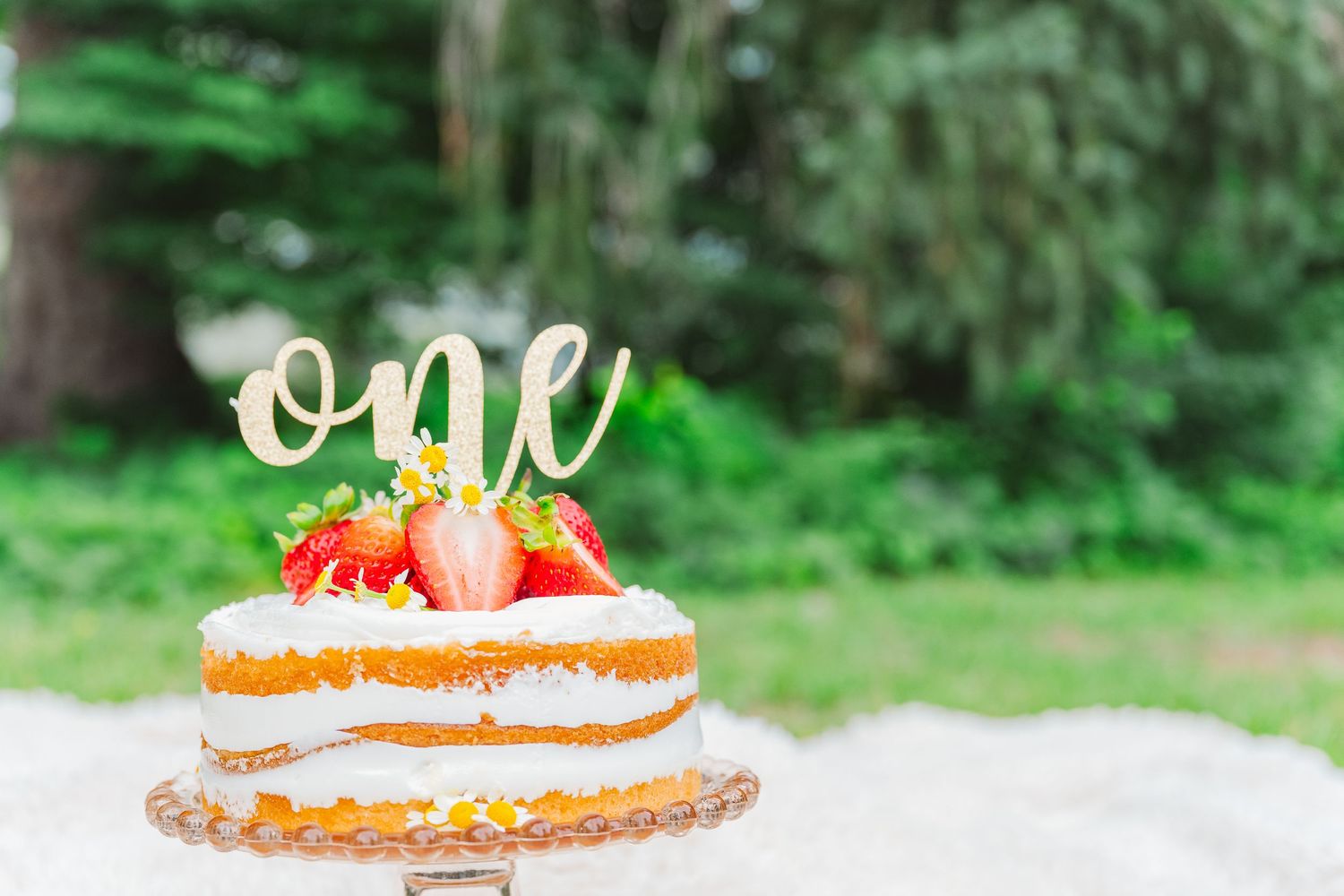Close up of white birthday cake with strawberries and gold 'one' topper against green foliage.