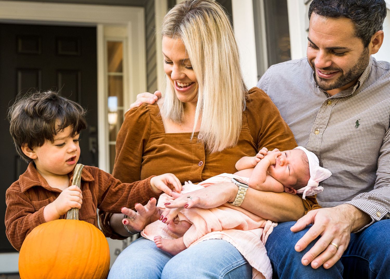 Family sitting together on a porch with a pumpkin and newborn baby sharing a tender moment.