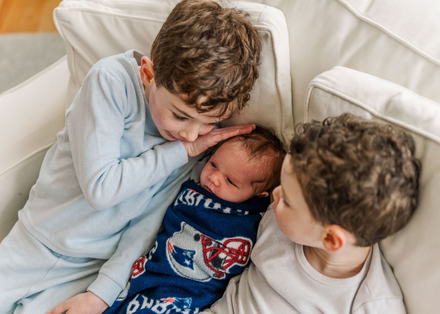 Two children snuggle close to baby sibling wearing blue pajamas on white bedding.