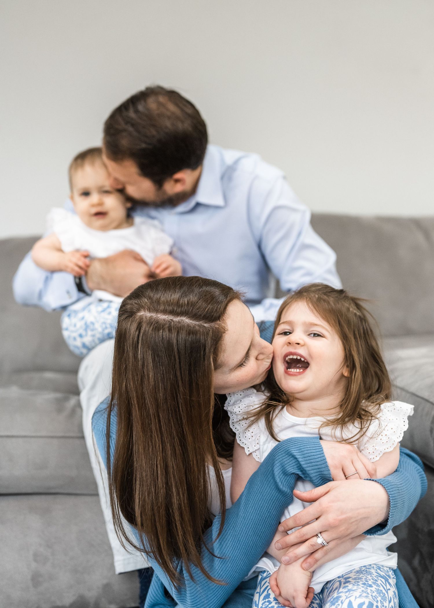 Playful family moment captured on gray couch with joyful expressions.