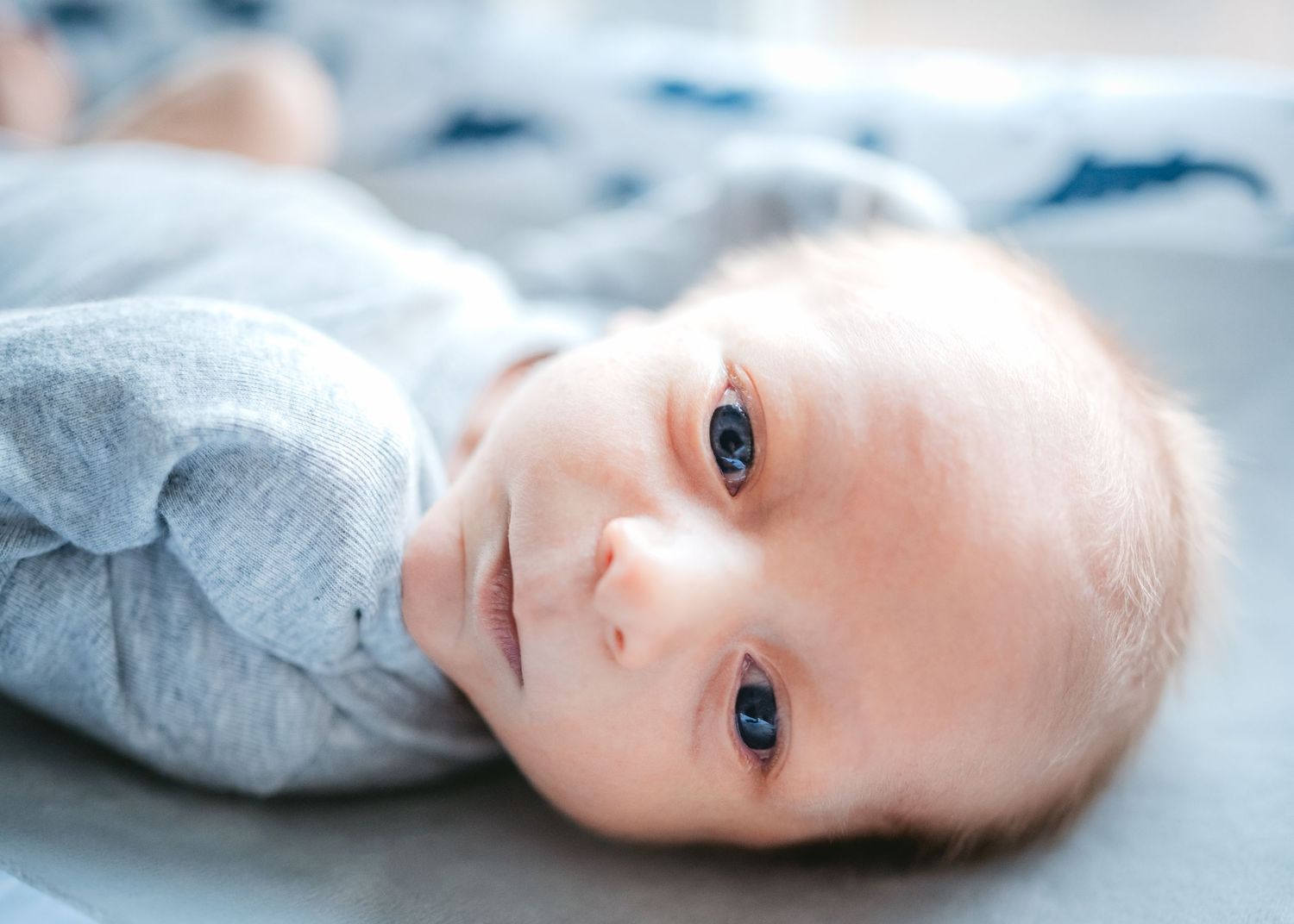 Baby with bright blue eyes lying on light colored bedding.