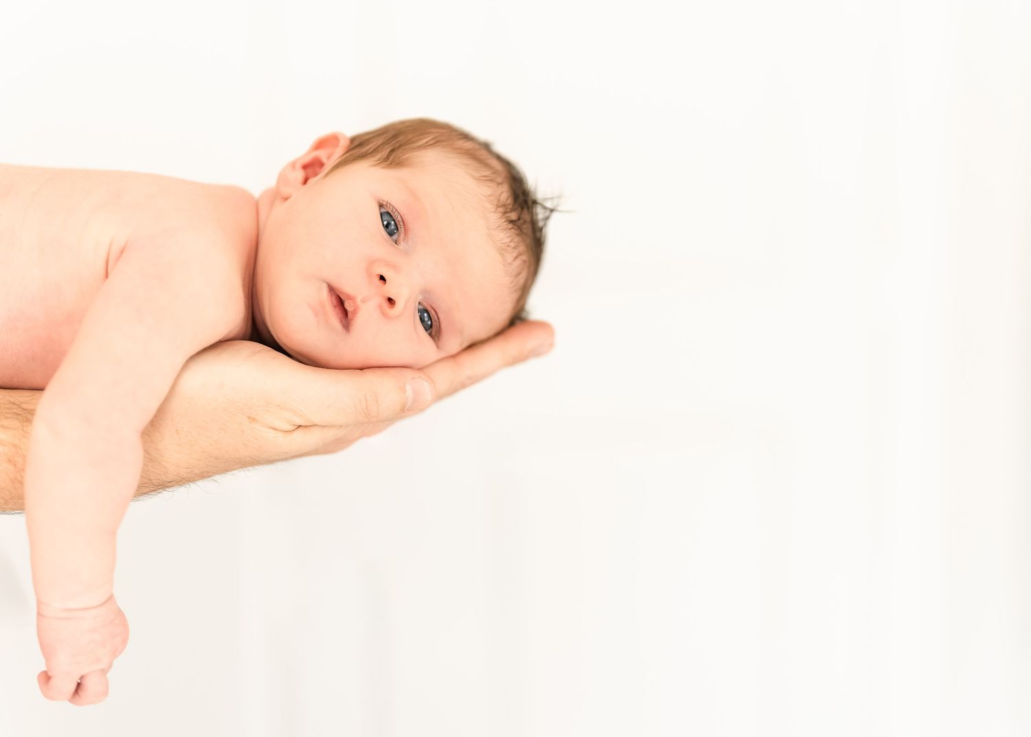 A baby lies on their side against a white surface, gazing softly at the camera.