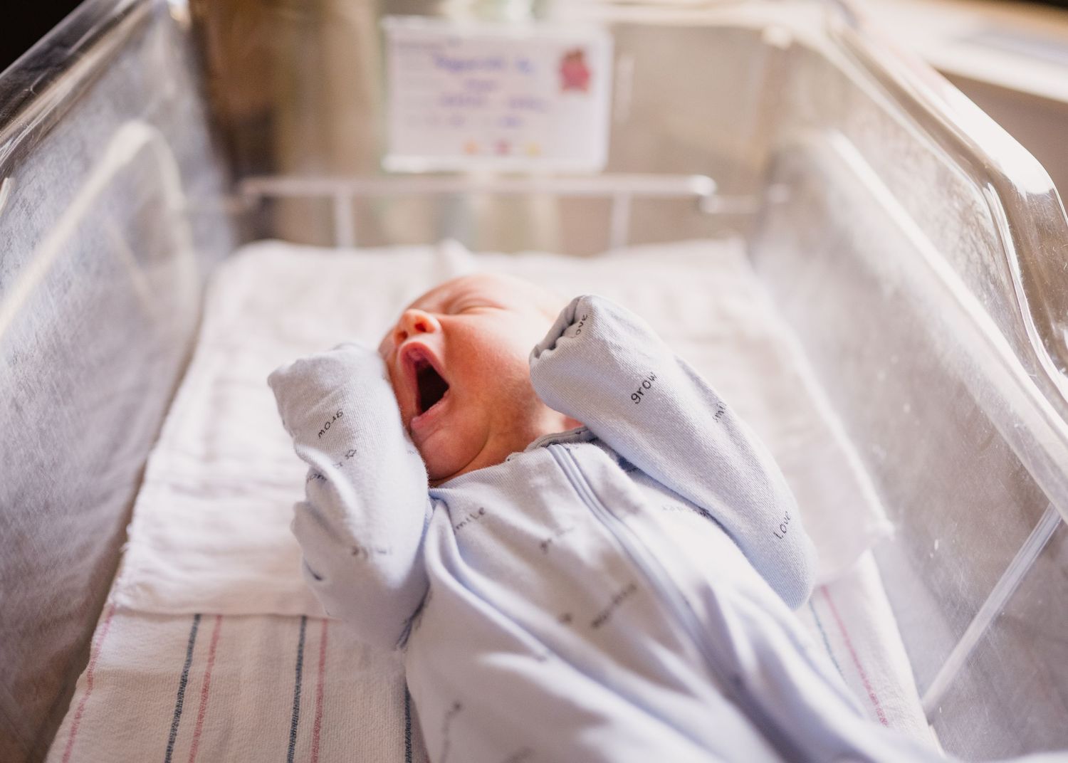 Newborn baby wearing white sleeper rests in a hospital bassinet.