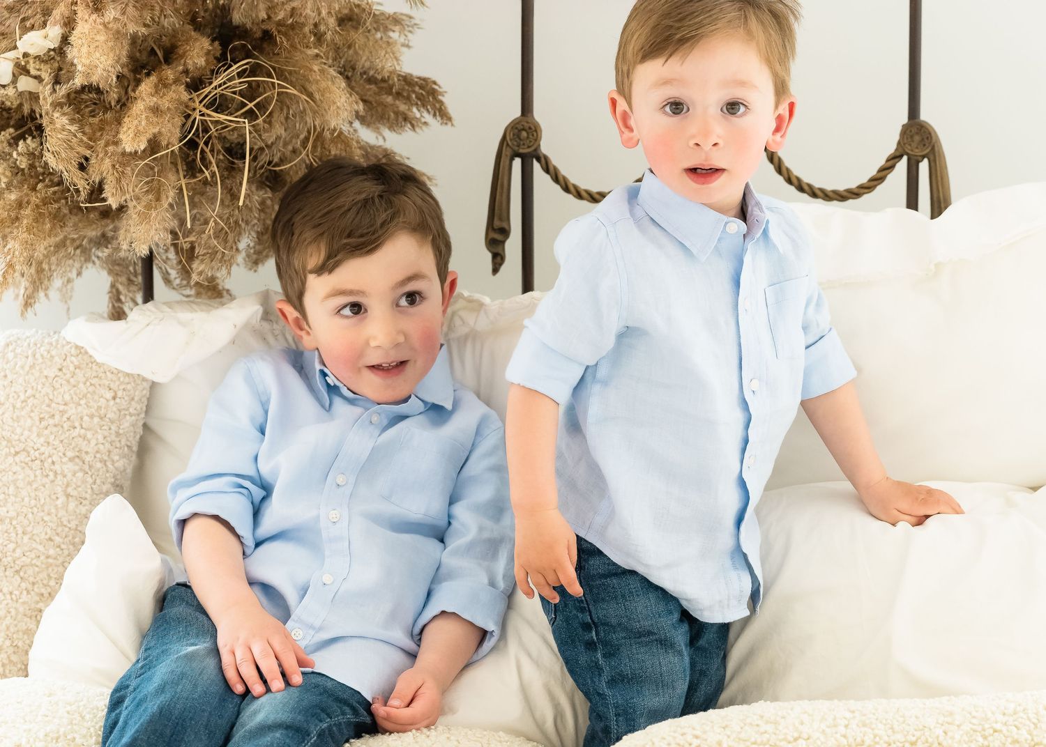 Two young children in matching light blue button-down shirts sit together on a white couch next to dried pampas grass.