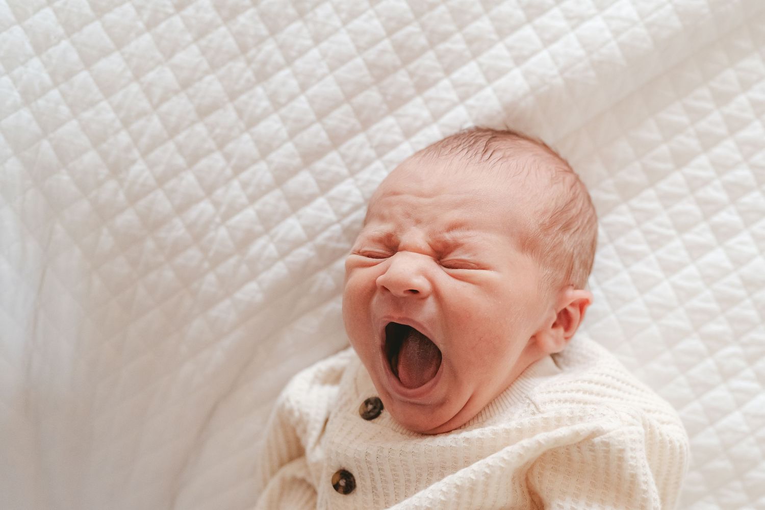 A newborn baby in white knit sweater yawning on white textured blanket.