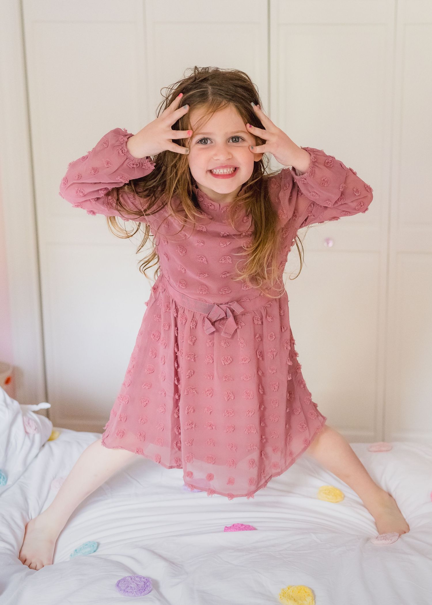 Child in pink dress playing and posing on white bed with colorful polka dots.