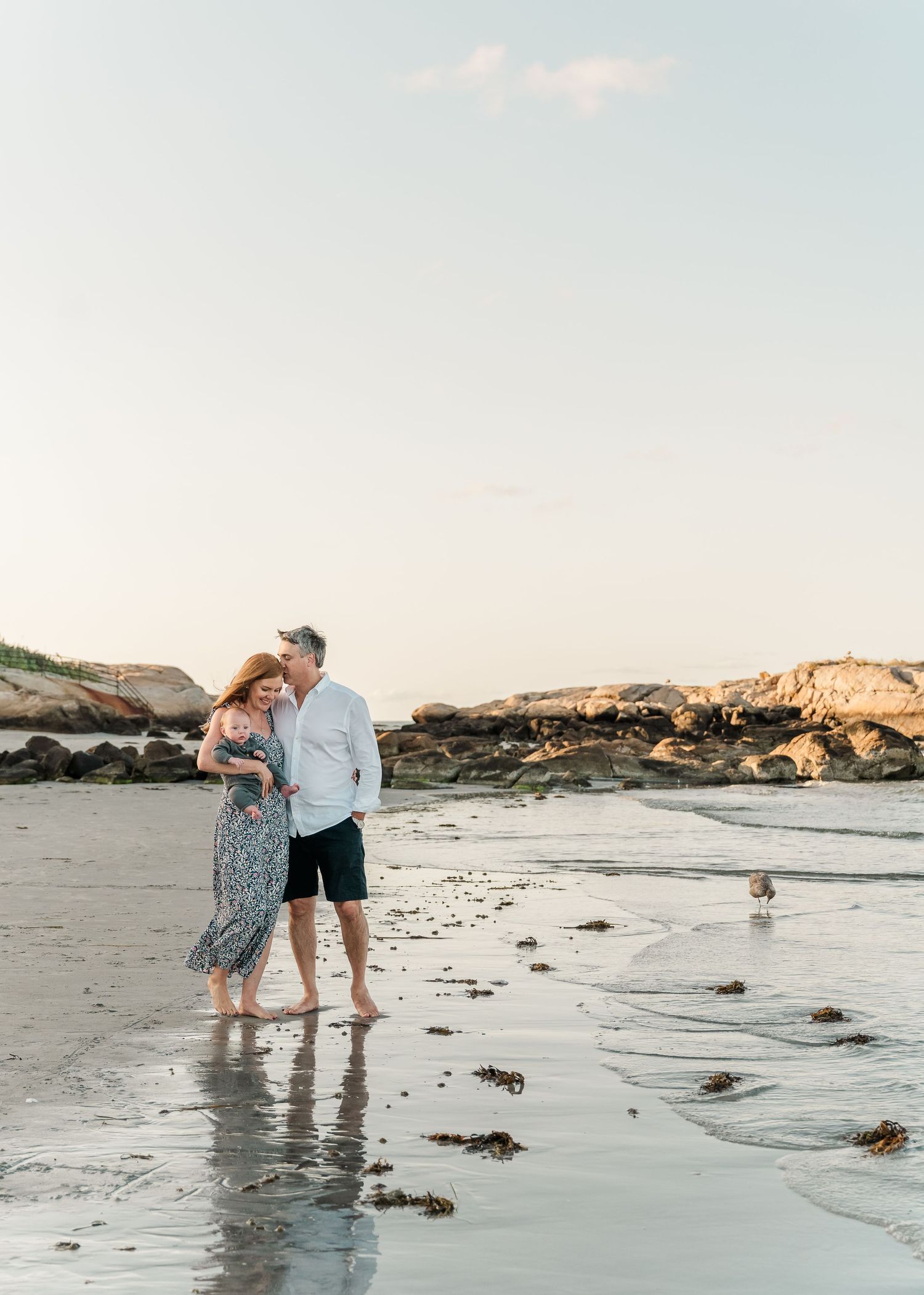 A couple walks together on a serene beach at sunset with rocky cliffs in the background.
