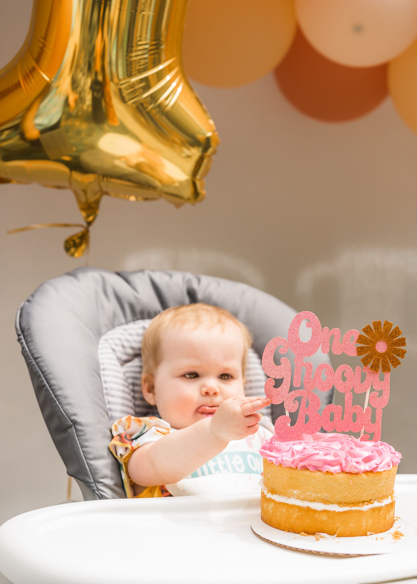 Baby in high chair with pink frosted birthday cake and gold balloon decoration.